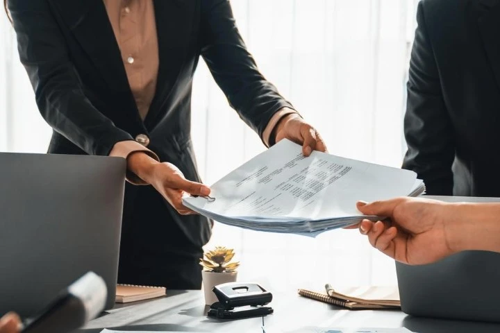 Two people standing by a desk handing over documents.